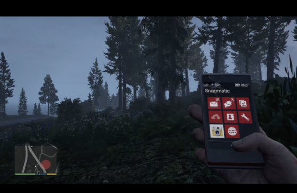 The image shows a screenshot from Grand Theft Auto V. From a first person perspective you look at a rural, forest area. The hand of your avatar holds a simulated smartphone showing a photo of the scene.